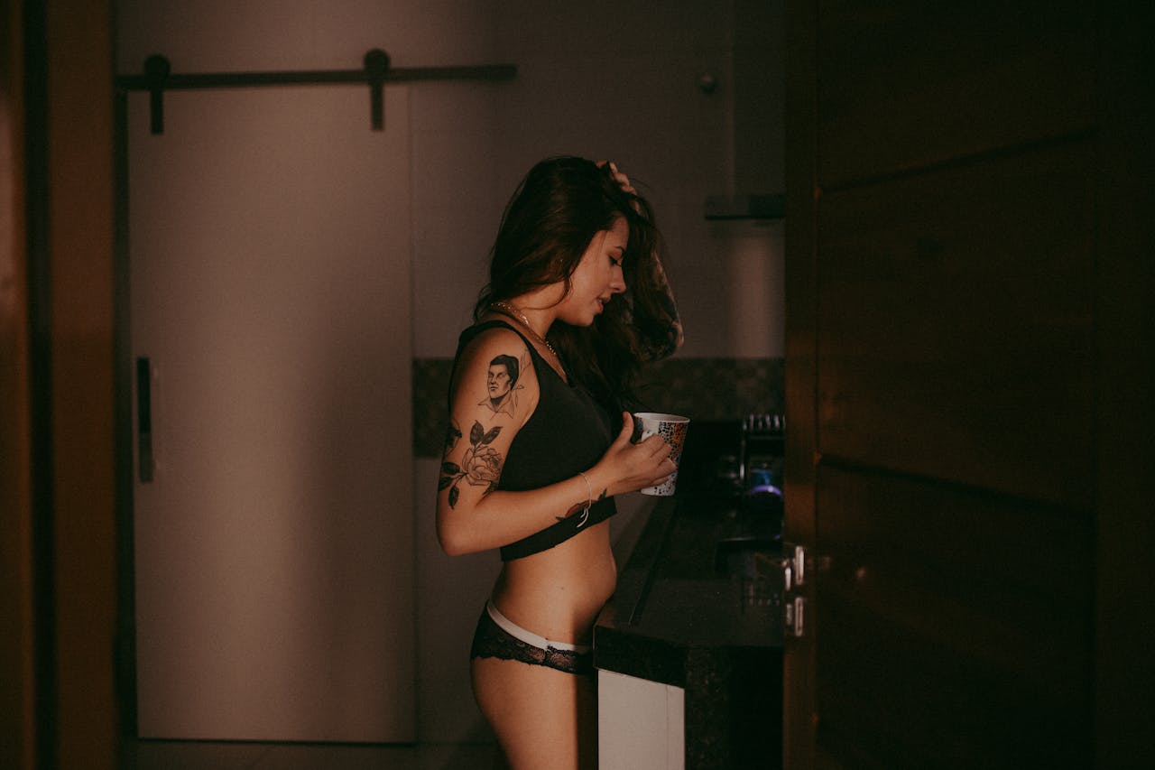 Facilities A young woman with tattoos, wearing lingerie, enjoys a casual moment indoors with a cup in hand.