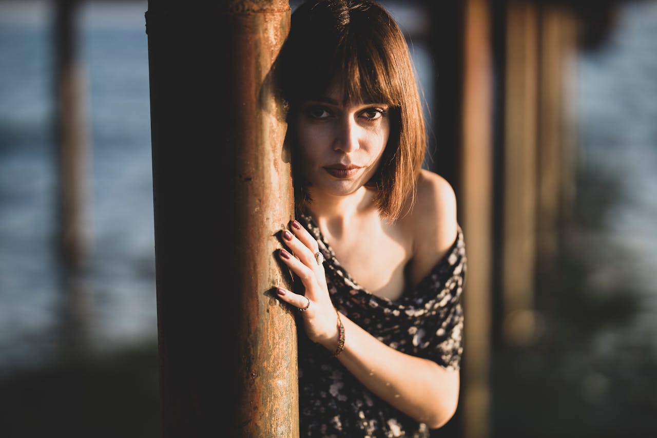 Stylish woman posing by a lakeside pier with soft lighting and fashion-forward attire.